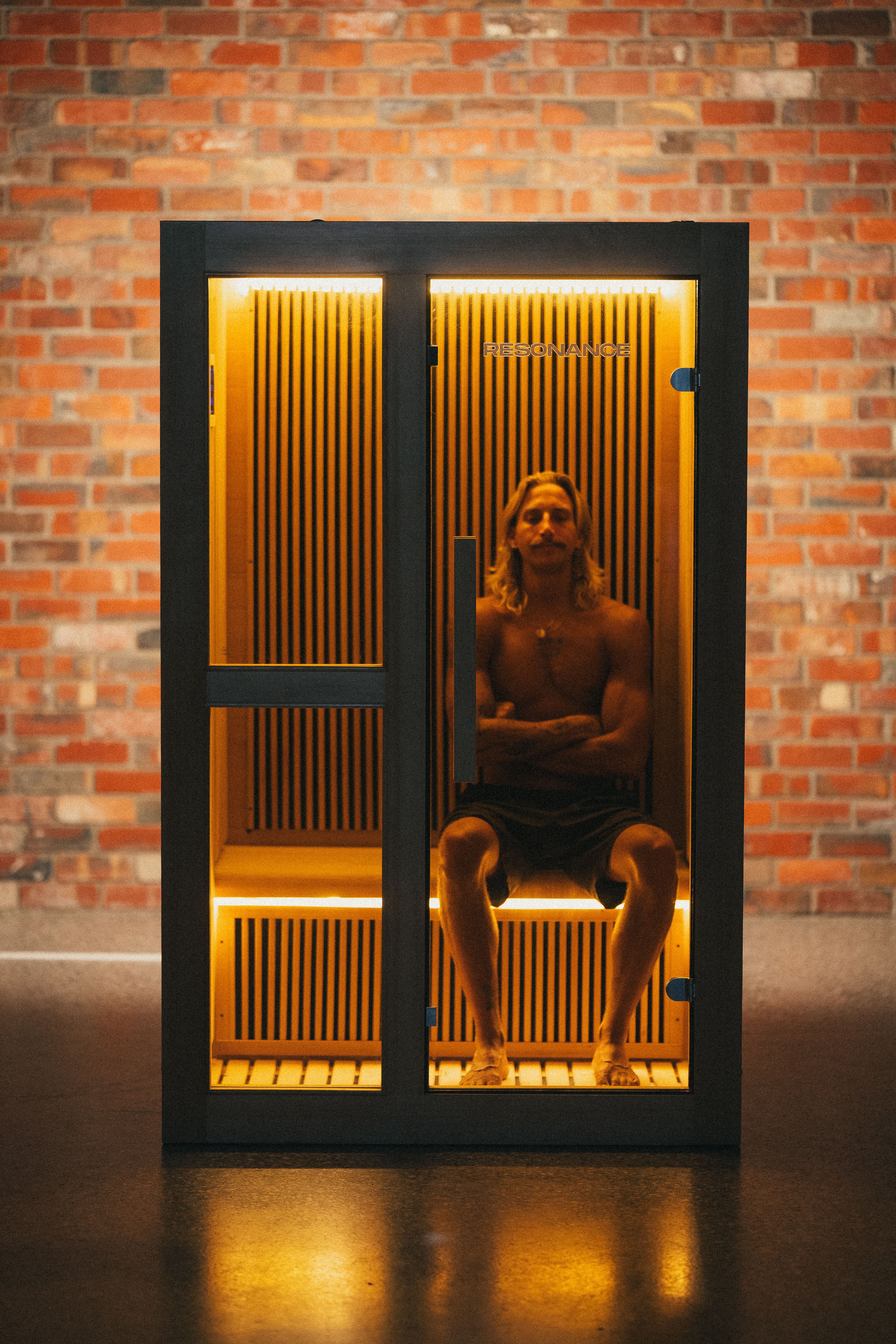 Athlete recovering inside Resonance Recovery Pod infrared sauna — 2-person cedar cabin with amber chromotherapy lighting and glass doors